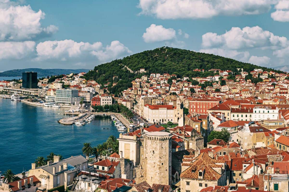 Blick von oben auf die kroatische Küstenstadt Split mit ihren historischen Steingebäuden, roten Ziegeldächern, dem Hafen und einem bewaldeten Hügel im Hintergrund.