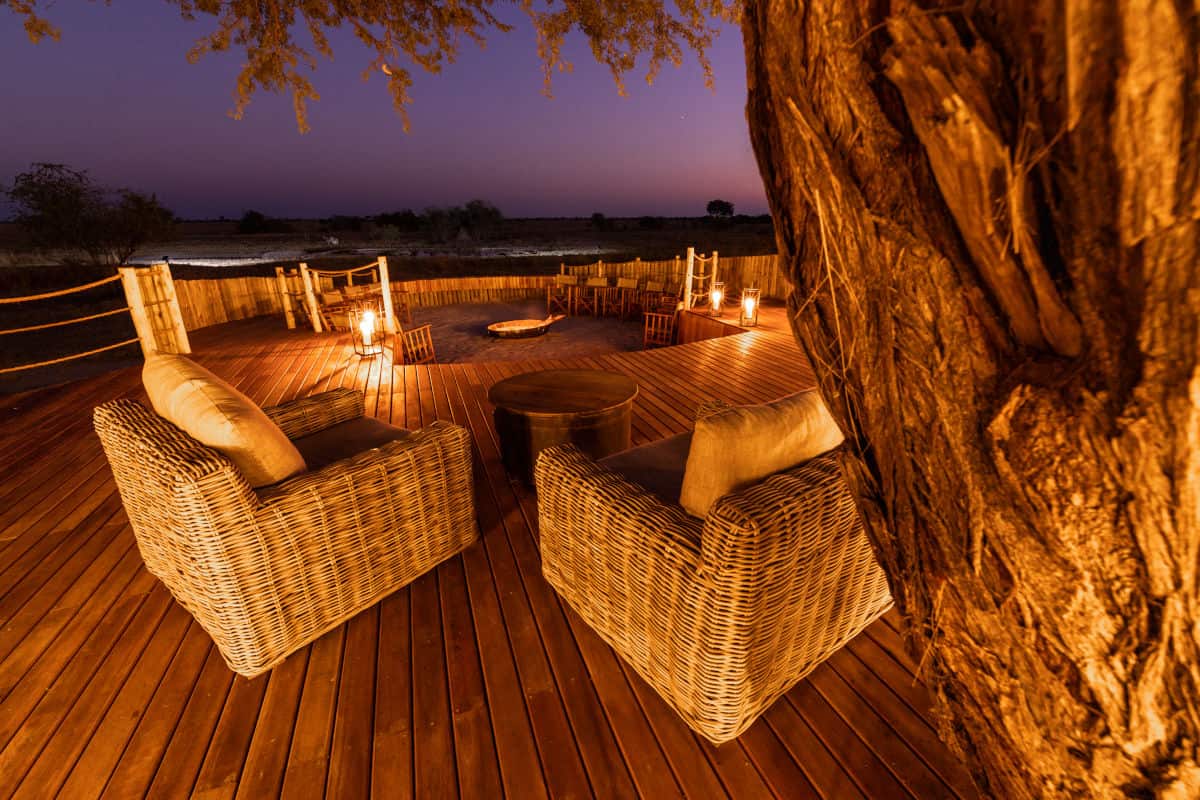 Zwei Korbsessel auf einer Holzterrasse mit Blick auf eine nächtliche Savannenlandschaft, beleuchtet durch eine Feuerstelle und warme Laternen im Freien.