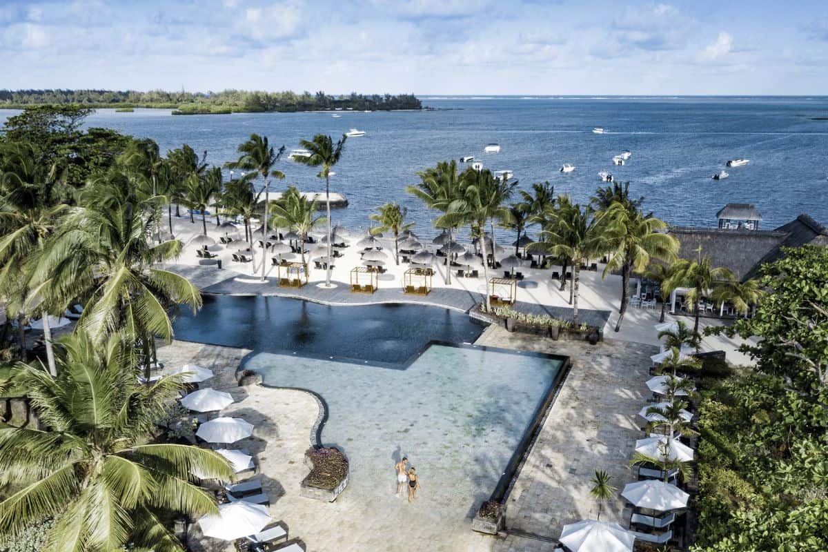 Luftaufnahme einer Hotelanlage mit einem großen, mehrteiligen Pool, Palmen, weißen Sonnenschirmen am Sandstrand und Booten auf dem blauen Meer im Hintergrund.