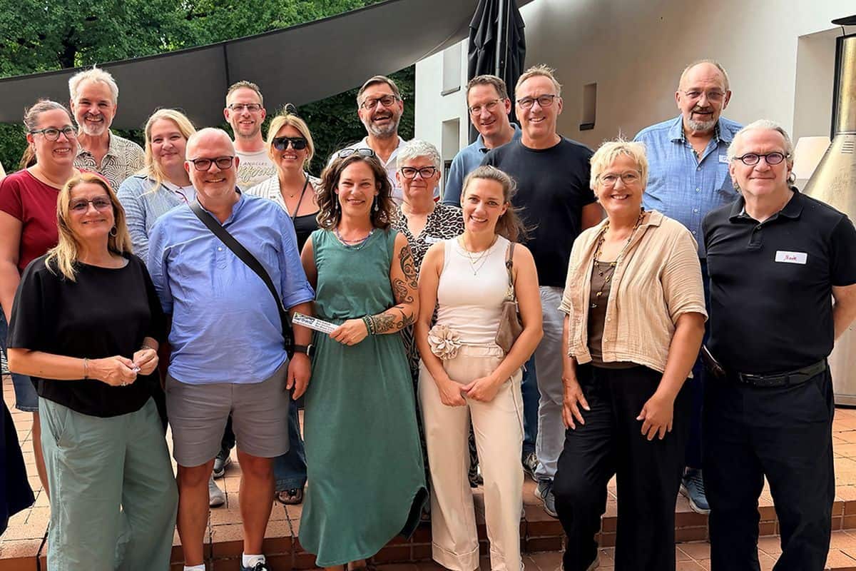 Eine Gruppe von 16 Personen unterschiedlichen Alters steht lächelnd auf einer Terrasse im Freien und blickt gemeinsam in die Kamera für ein Gruppenfoto.