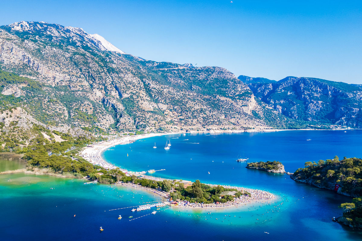 Luftaufnahme der Bucht von Ölüdeniz in der Türkei mit einer sichelförmigen Landzunge, türkisblauem Wasser, bewaldeten Bergen und mehreren kleinen Booten.