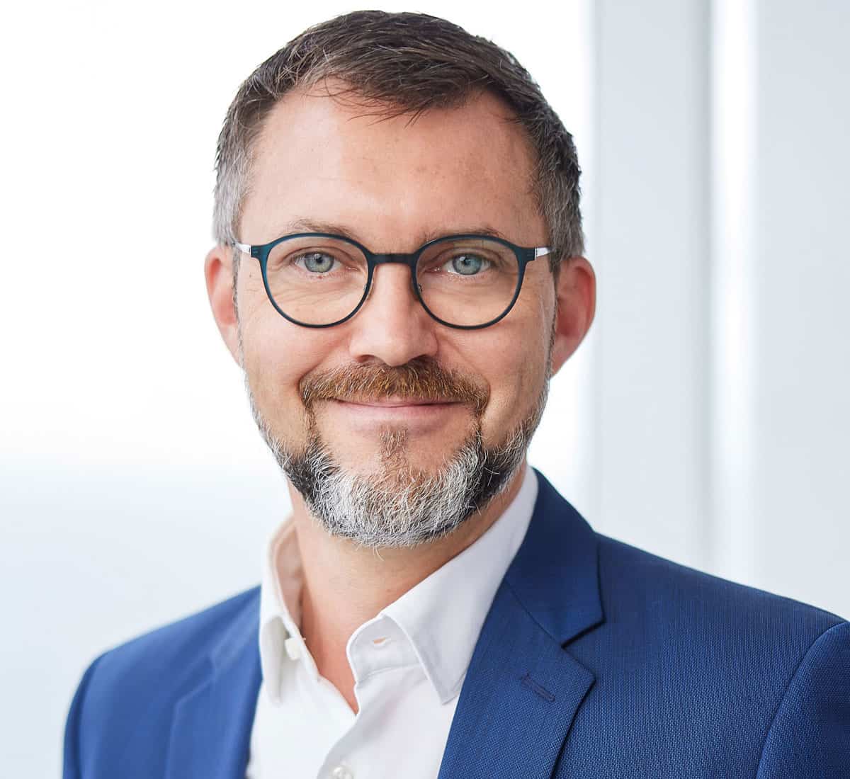 Ein Mann mit Brille, kurzem Haar und Bart trägt ein weißes Hemd sowie ein blaues Sakko und blickt freundlich lächelnd direkt in die Kamera des Porträts.