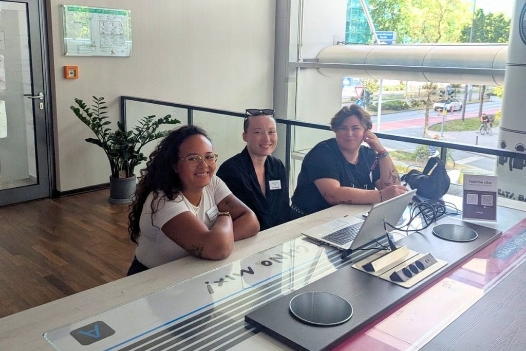 Drei Frauen sitzen lächelnd an einem hellen Schreibtisch mit Laptop in einem modernen Bürogebäude mit großen Fensterfronten und Blick auf eine Straße.