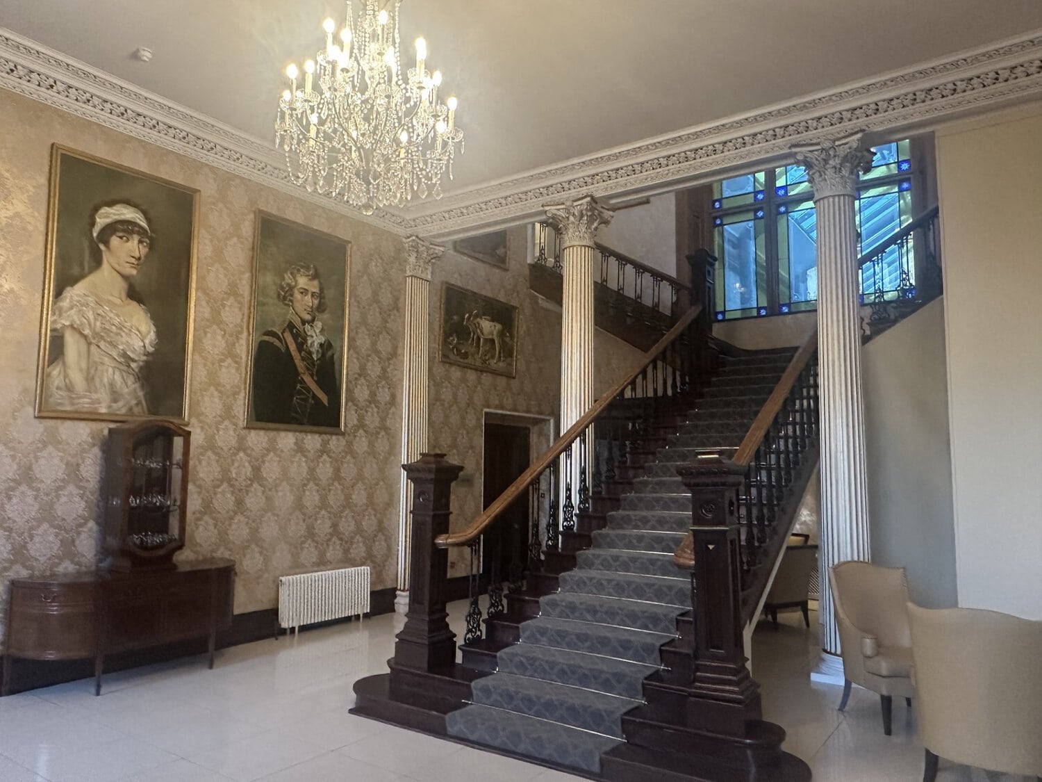 Ein prunkvolles Foyer mit einer großen Holztreppe, antiken Ölgemälden an der gemusterten Wand, einem gläsernen Kronleuchter und klassischen Säulen im Raum.