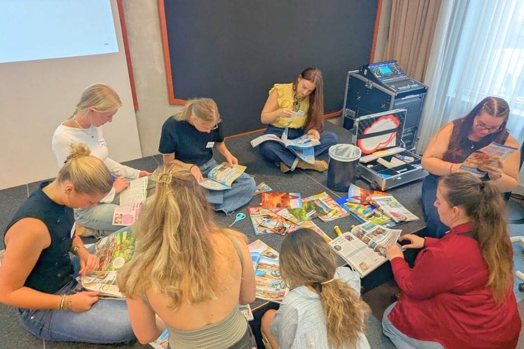 Eine Gruppe von acht jungen Frauen sitzt auf einem Boden in einem Raum im Kreis und betrachtet konzentriert verschiedene aufgeschlagene Zeitschriften.