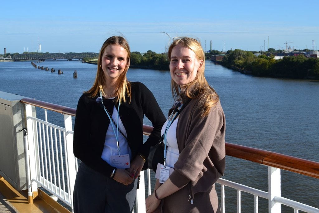 Zwei junge Frauen mit Namensschildern an Umhängebändern stehen lächelnd an der Reling eines Schiffes vor einer Flusslandschaft mit Brücke im Hintergrund.