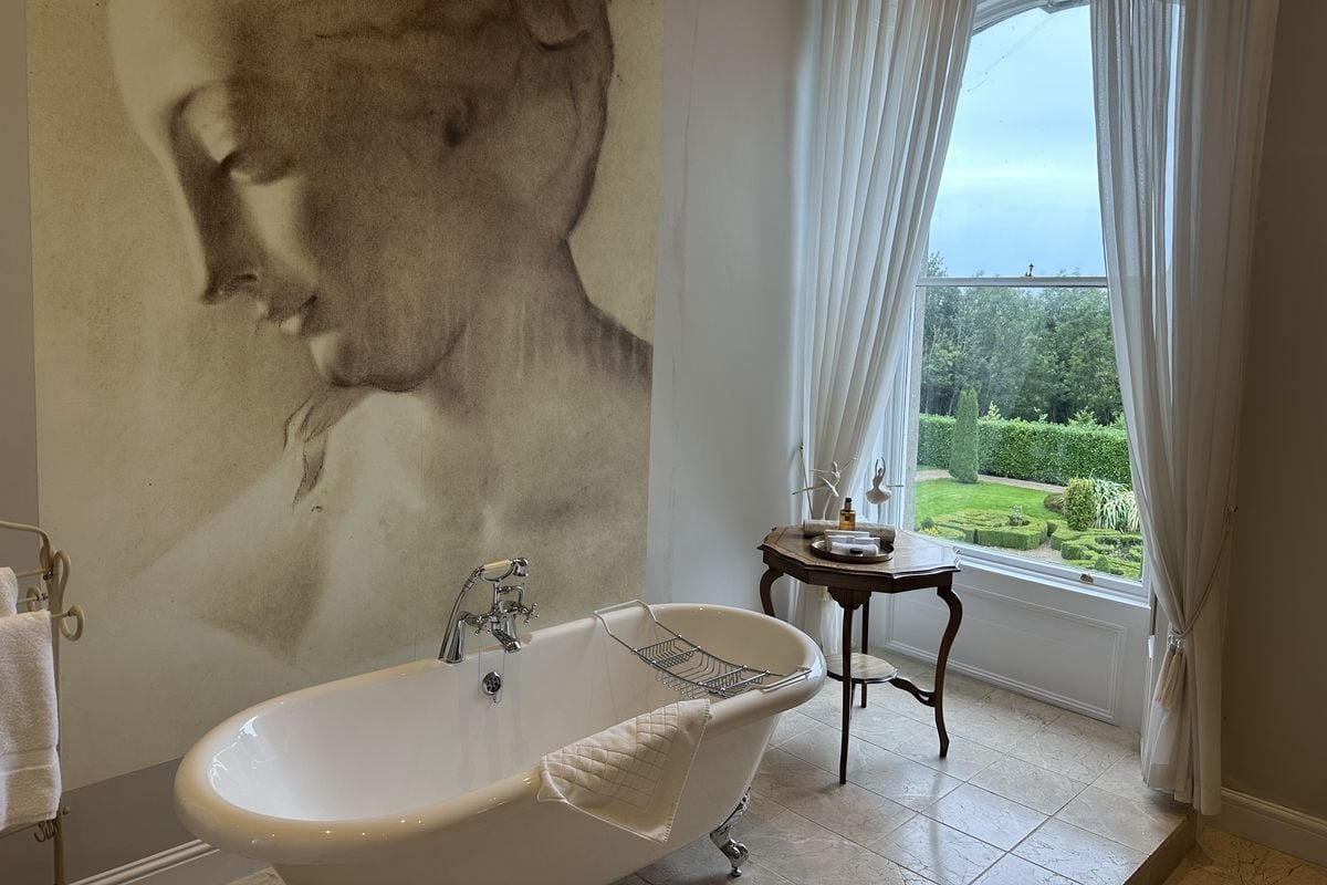 Ein Badezimmer mit einer freistehenden Badewanne vor einer Wand mit großem Porträt-Wandbild und einem Fenster mit Blick auf einen gepflegten Garten.