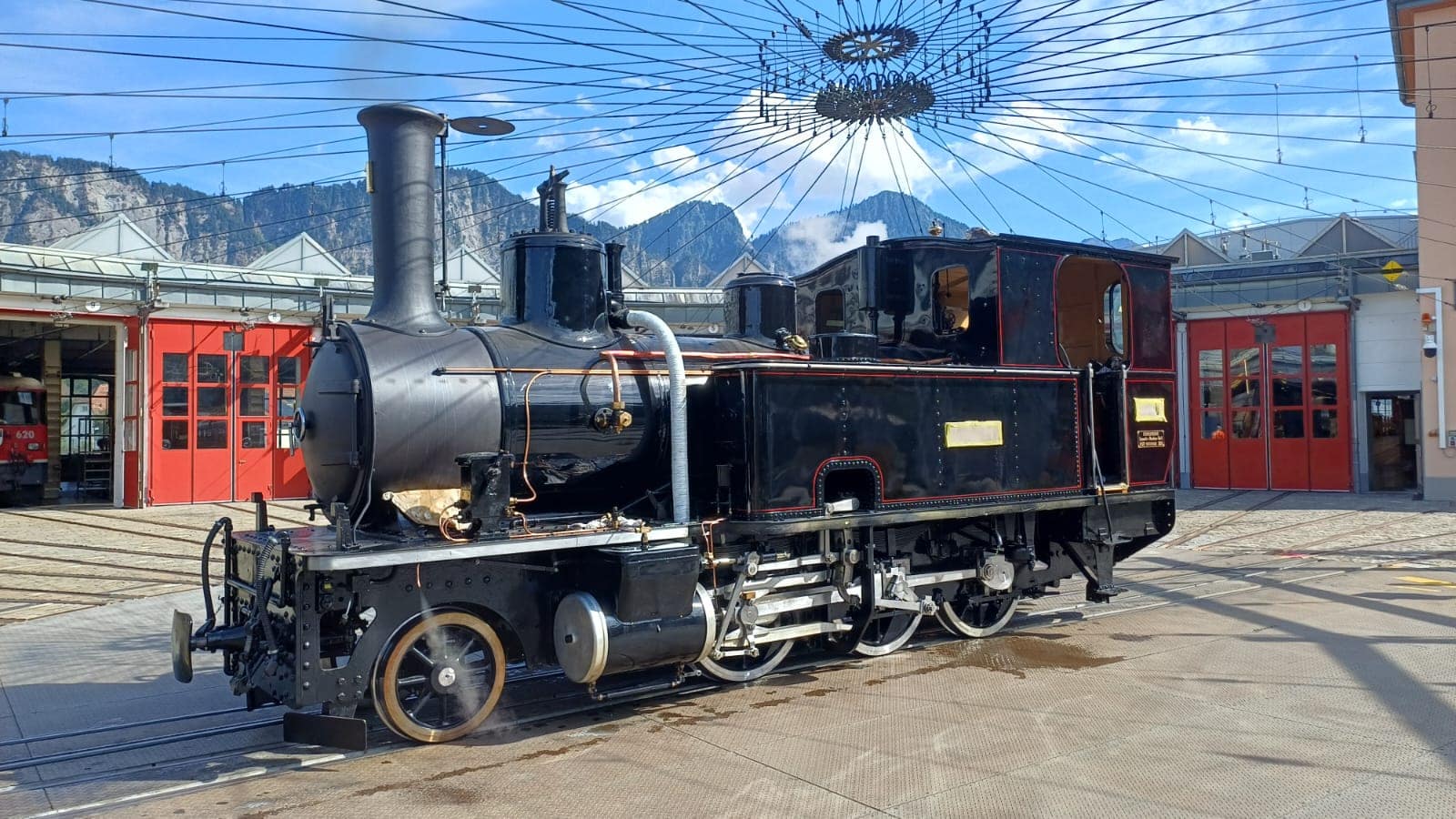 Eine historische schwarze Dampflokomotive steht auf einem Gleis vor einem Depot mit roten Toren, im Hintergrund sind Berge unter blauem Himmel zu sehen.
