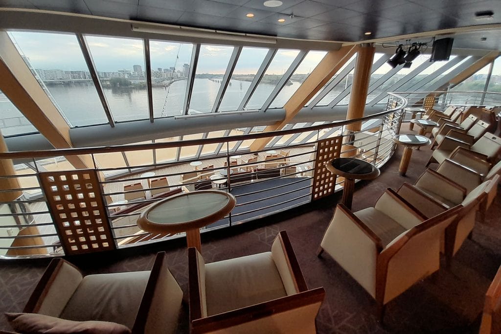 Blick aus einer Lounge mit großen Glasfronten auf einen Fluss und eine Stadt am Ufer, davor stehen beige Sessel und kleine Tische auf einem Teppichboden.