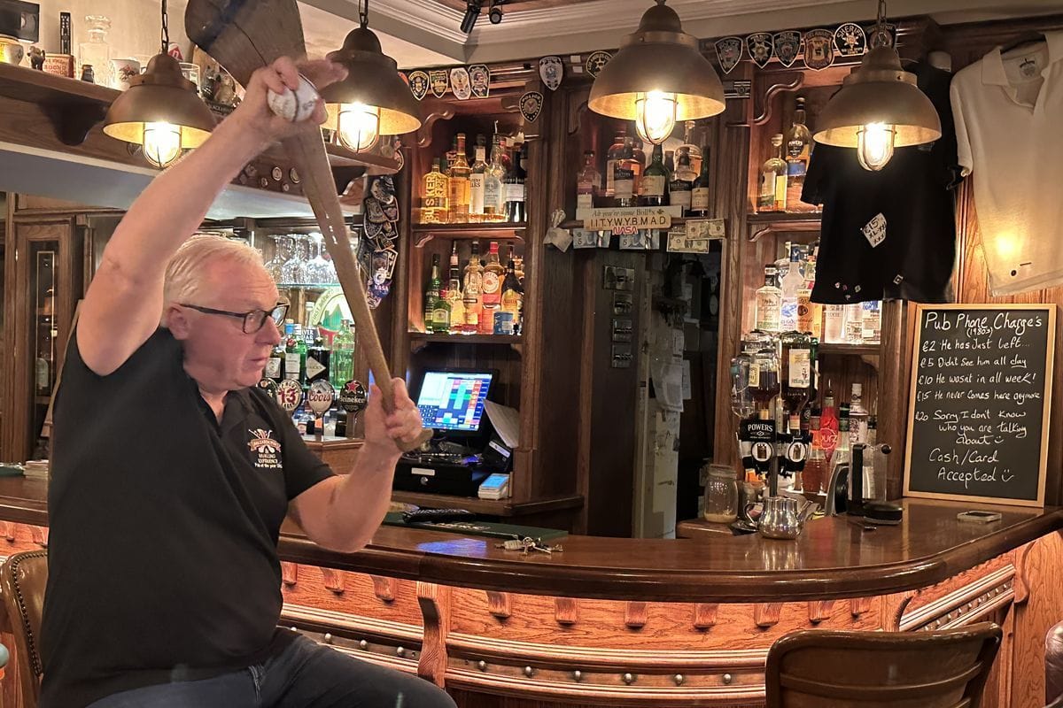 Ein Mann mit Brille sitzt an einer hölzernen Pub-Theke und hält ein langes, hölzernes Sportgerät mit einem Ball in der Hand in die Höhe vor dem Tresen.