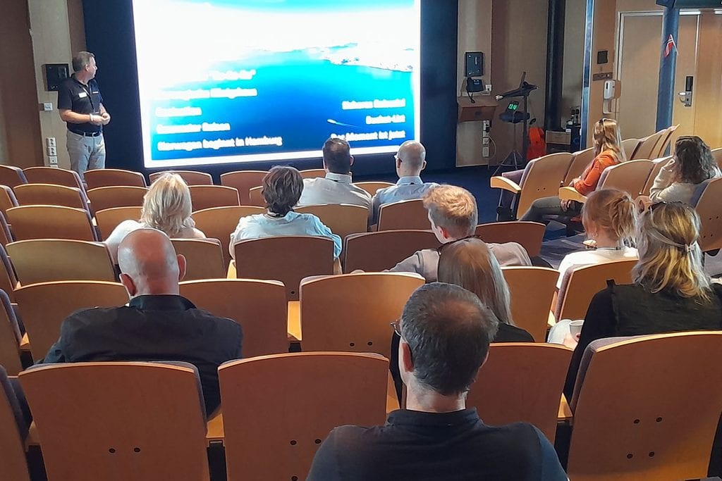 Ein Mann steht vor einer großen Leinwand in einem Konferenzraum und hält einen Vortrag vor einem Publikum, das in Reihen von Holzstühlen sitzt.