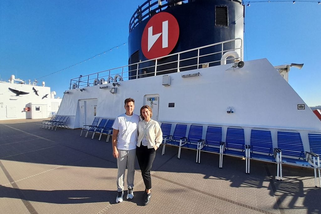 Ein junger Mann und eine junge Frau stehen nebeneinander auf dem Außendeck eines Schiffes vor einem großen schwarzen Schornstein mit rotem H-Logo.