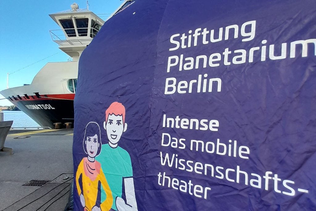 Ein großes, blaues Banner der Stiftung Planetarium Berlin mit Comicfiguren steht vor dem Bug eines Schiffes mit der Aufschrift Midnatsol am Kai.