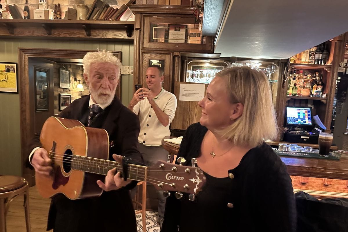 Ein älterer Mann mit weißem Bart spielt Gitarre in einem Pub, während eine Frau ihn anlächelt und ein Mann im Hintergrund die Szene mit dem Handy filmt.