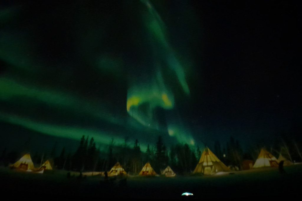 Grüne Nordlichter leuchten am dunklen Nachthimmel über einer Ansammlung von beleuchteten Tipis, die vor einer dunklen Waldkulisse in einer Ebene stehen.