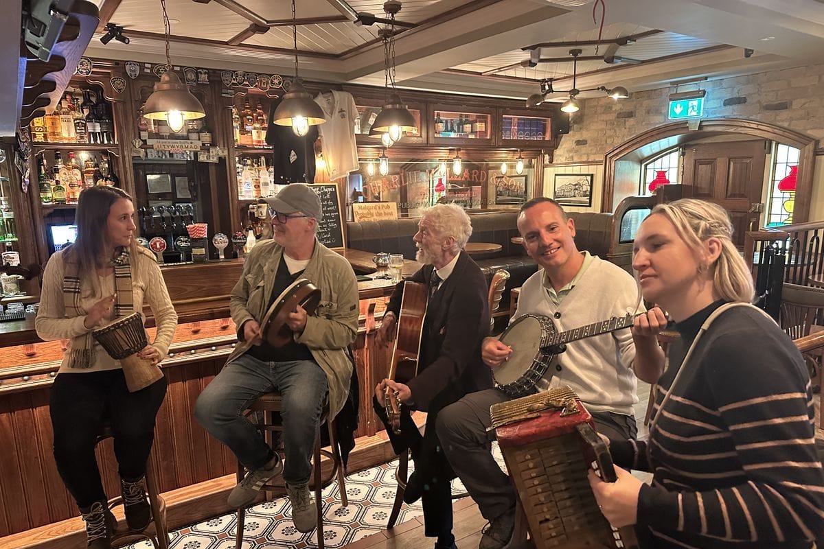 Eine Gruppe von fünf Personen musiziert gemeinsam in einem traditionellen Pub mit Holztresen, Instrumenten wie Gitarre, Banjo, Akkordeon und Trommel.