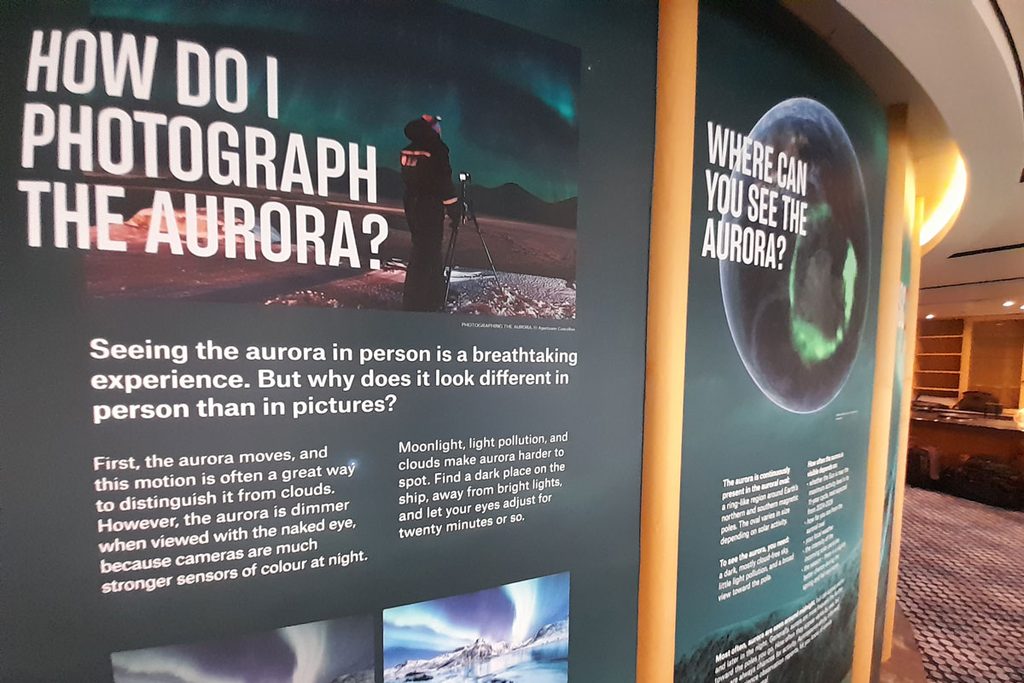 Eine dunkelgrüne Informationstafel mit englischen Texten und Fotos über Polarlichter, montiert an einer geschwungenen Wand in einem Innenraum mit Teppich.
