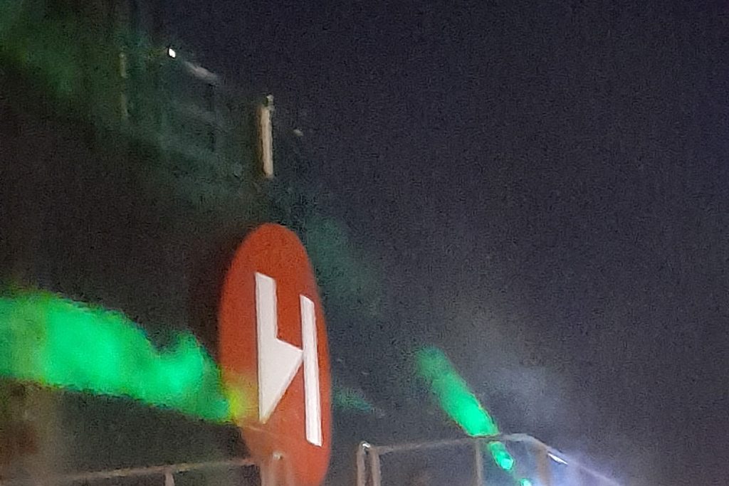 Nahaufnahme eines roten Logos mit einem weißen Buchstaben H auf einem dunklen Schiffskörper bei Nacht, beleuchtet durch grünes Licht und leichten Nebel.