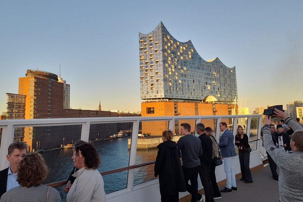 Blick von einem Schiffsdeck auf die Hamburger Elbphilharmonie bei Sonnenuntergang, während mehrere Personen am Geländer stehen und die Aussicht genießen.