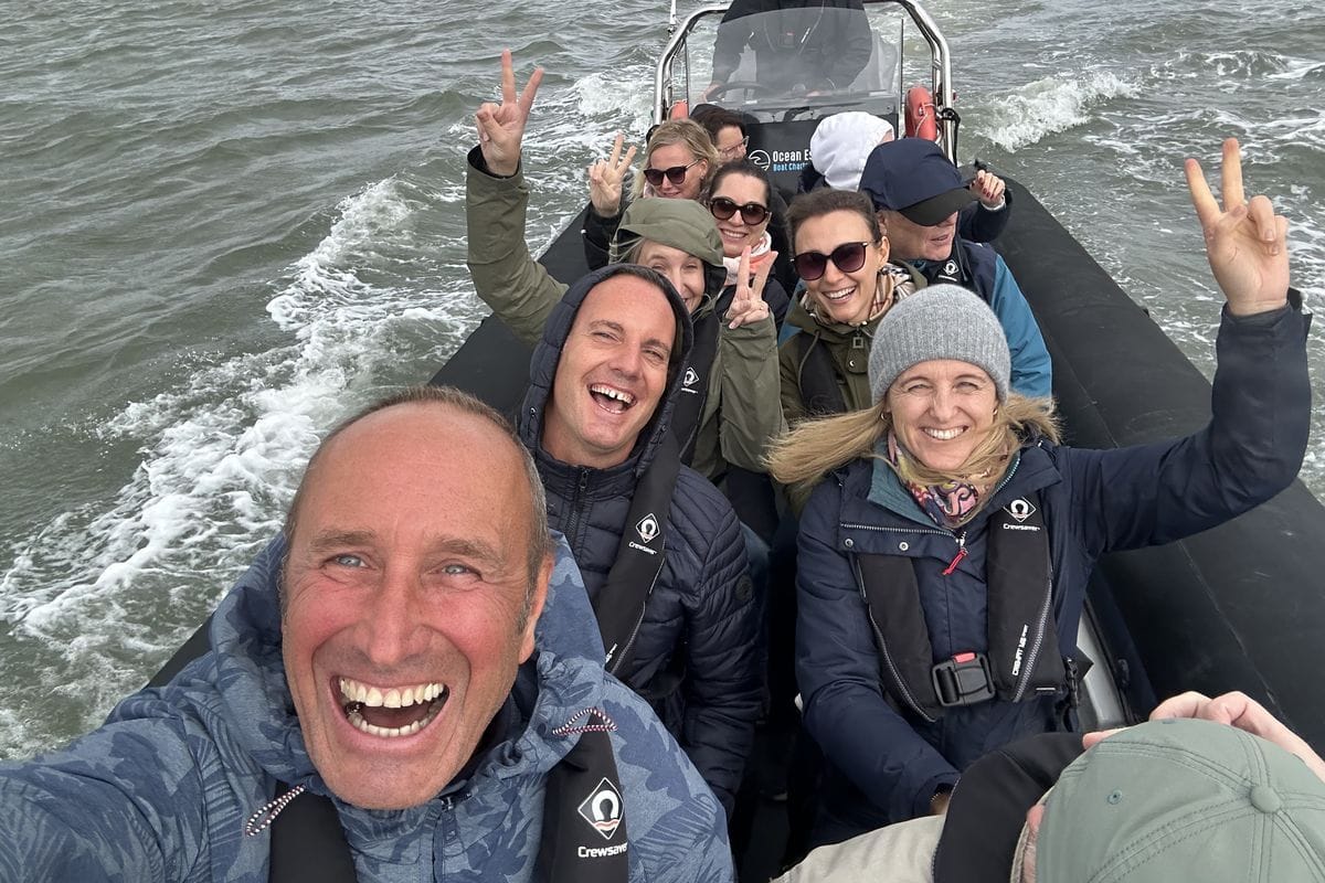 Eine Gruppe von Menschen in Outdoor-Kleidung und Schwimmwesten macht ein Selfie auf einem schnell fahrenden Schlauchboot auf dem bewegten, grauen Wasser.