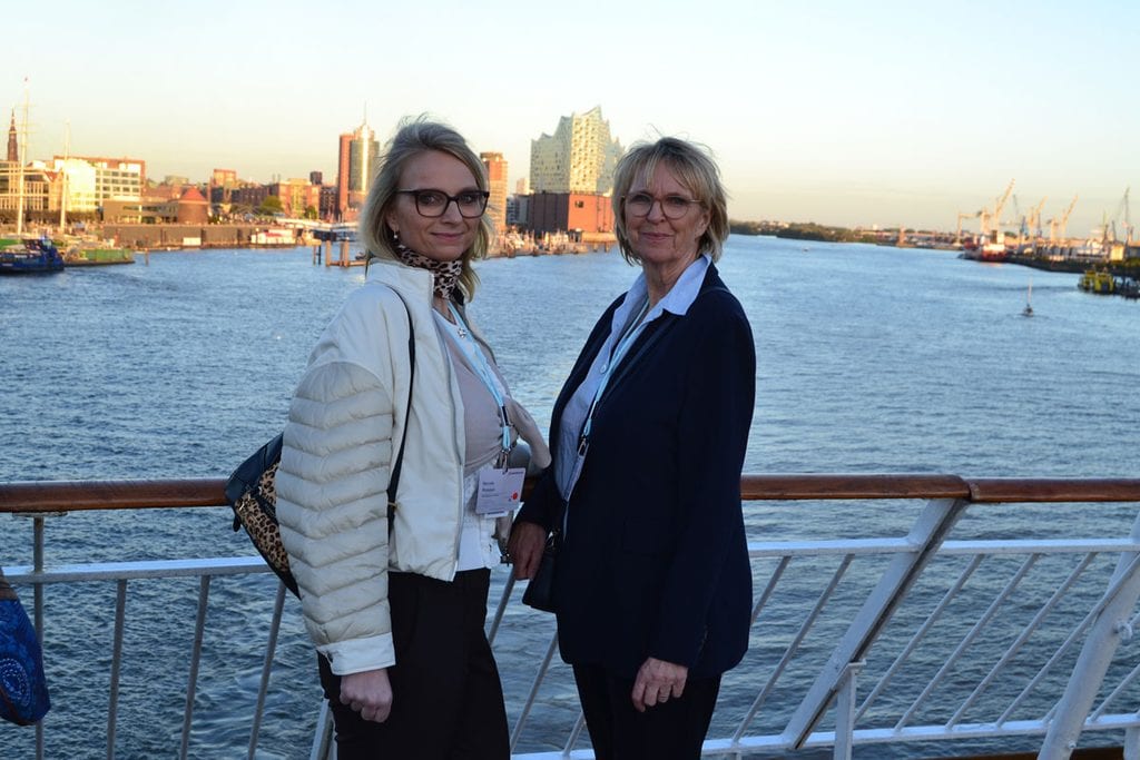 Zwei Frauen stehen an einer Reling vor der Hamburger Hafenkulisse mit der Elbphilharmonie im Hintergrund und blicken in die Kamera des Fotografen.