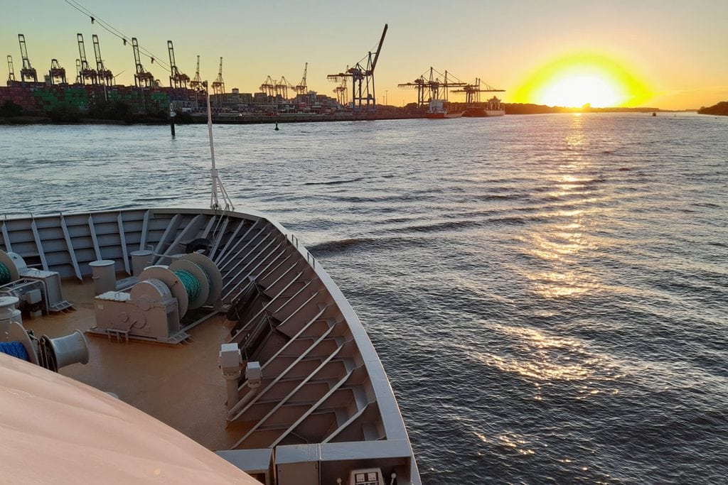 Blick vom Bug eines Schiffes auf einen Containerhafen mit Kränen bei Sonnenuntergang, wobei sich das Licht auf der Wasseroberfläche des Flusses spiegelt.