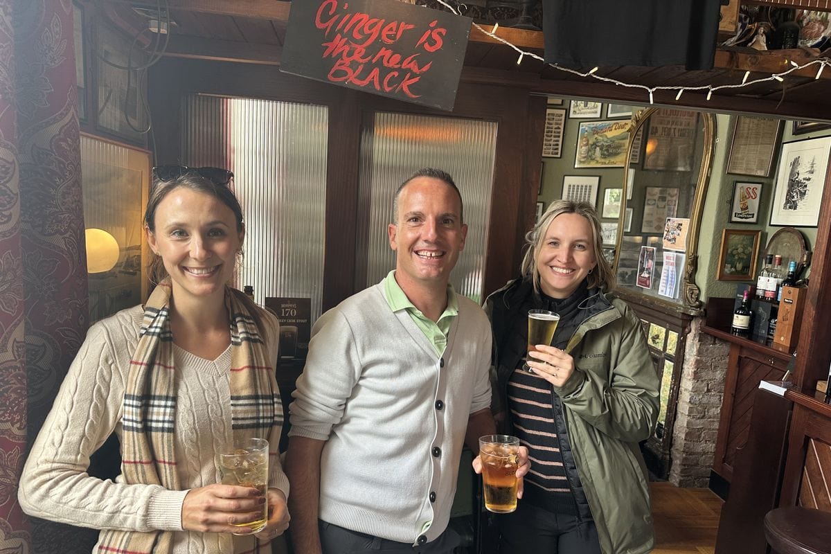 Zwei Frauen und ein Mann stehen lächelnd in einem traditionellen Pub mit gerahmten Bildern an der Wand und halten jeweils ein Glas mit einem Getränk in Hand.