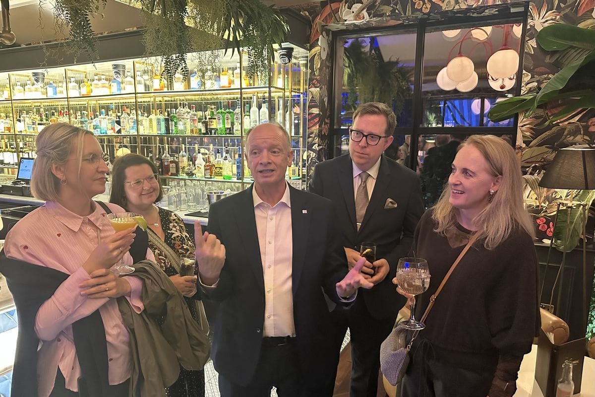 Eine Gruppe von vier Personen steht in einer Bar vor einem beleuchteten Regal mit Spirituosen und unterhält sich, während sie Gläser in den Händen halten.