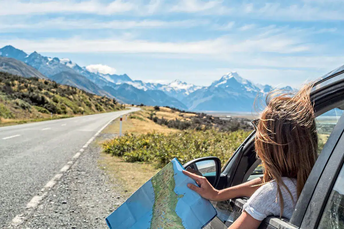 Eine Frau mit wehendem Haar schaut aus dem Fenster eines Autos und hält eine geöffnete Landkarte vor einer weiten Berglandschaft mit einer leeren Straße.