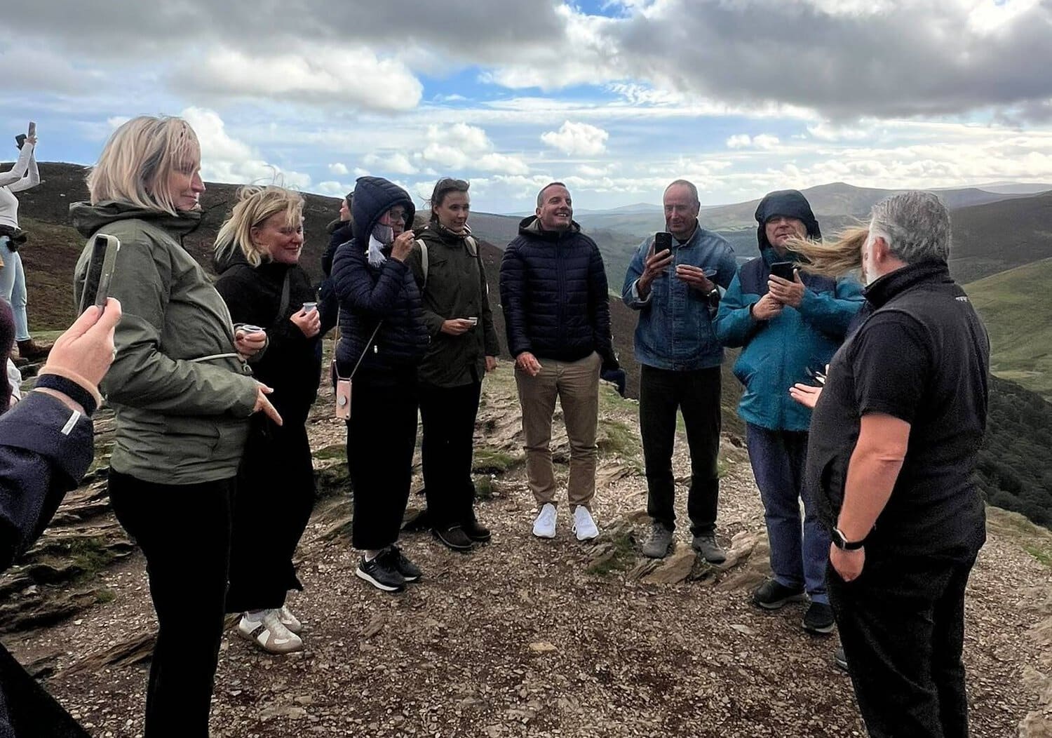 Eine Gruppe von Menschen steht auf einem hügeligen Pfad in einer bergigen Landschaft und hält kleine Becher oder Smartphones in den Händen.