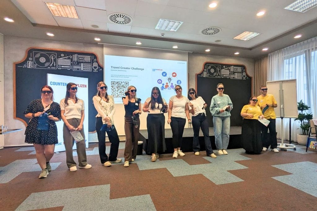 Eine Gruppe von neun Personen steht in einem Konferenzraum vor einer Leinwand mit QR-Code und trägt dunkle Sonnenbrillen, während sie in die Kamera blickt.
