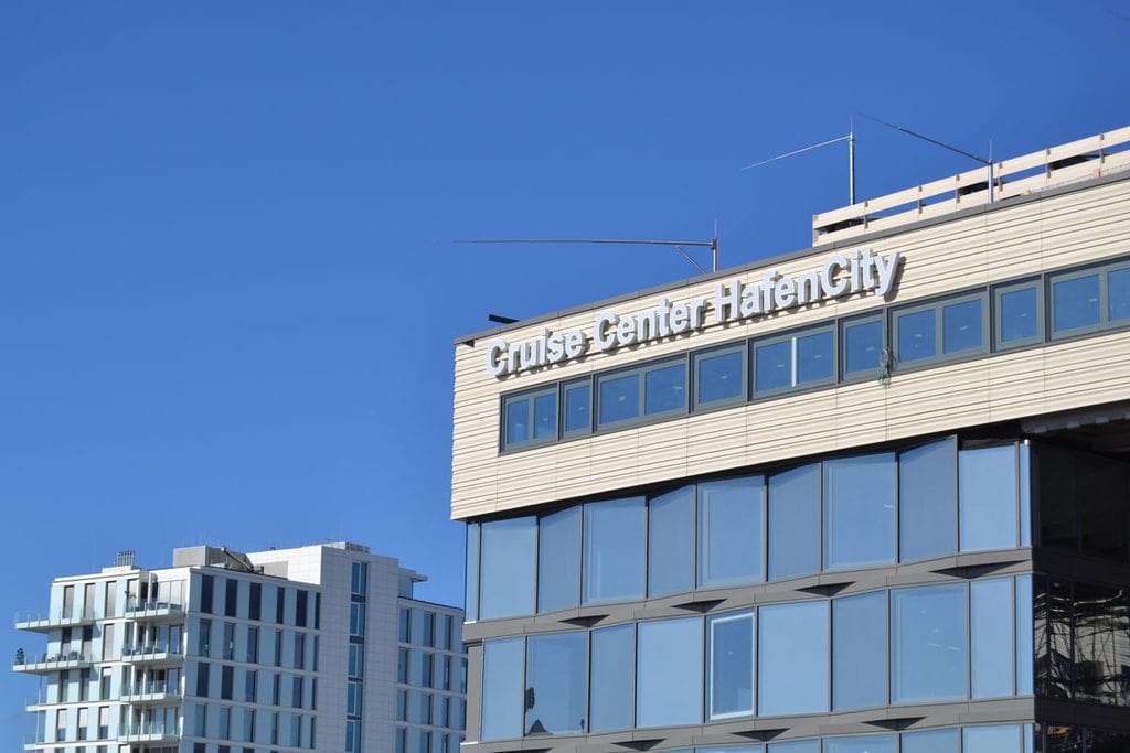 Die Fassade des Cruise Center HafenCity in Hamburg mit dem Schriftzug auf dem Dach vor einem klaren, blauen Himmel und einem weiteren Gebäude im Hintergrund.