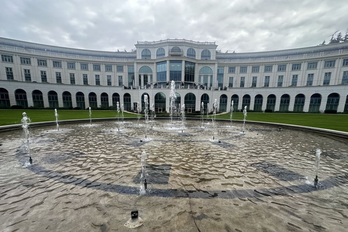 Ein großes, weißes, halbkreisförmiges Gebäude mit vielen Fenstern und Arkaden steht hinter einem runden Springbrunnen mit zahlreichen aufsteigenden Fontänen.