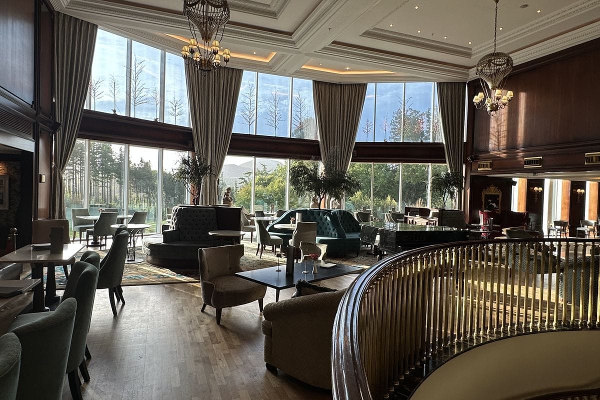 Ein eleganter Hotel-Loungebereich mit bodentiefen Fenstern, gepolsterten Sesseln, Sofas, kleinen Tischen und einem geschwungenen Geländer im Vordergrund.