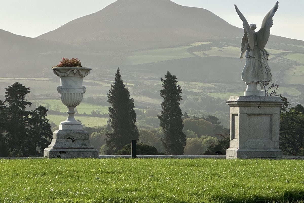 Eine steinerne Engelsstatue und eine dekorative Vase auf Sockeln stehen auf einer grünen Wiese vor einer hügeligen Landschaft mit Bäumen im Hintergrund.