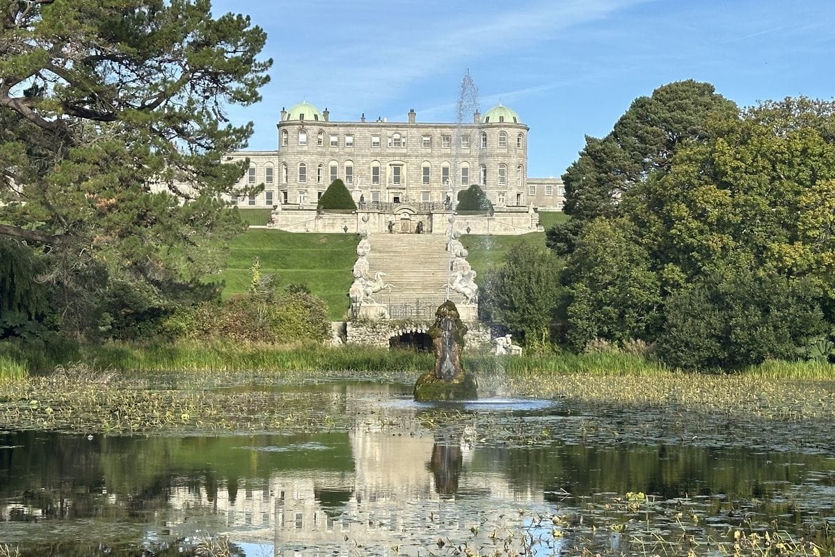 Ein großes, klassizistisches Herrenhaus mit zwei Kuppeln steht hinter einer breiten Steintreppe, die zu einem Teich mit einer sprudelnden Fontäne hinabführt.