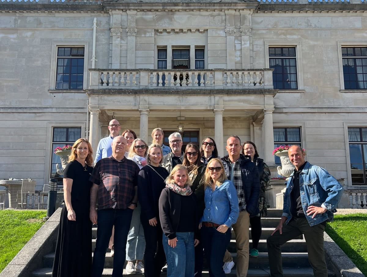 Eine Gruppe von 14 Personen steht auf einer breiten Steintreppe vor einem historischen Herrenhaus mit klassizistischer Fassade und einem Balkon.