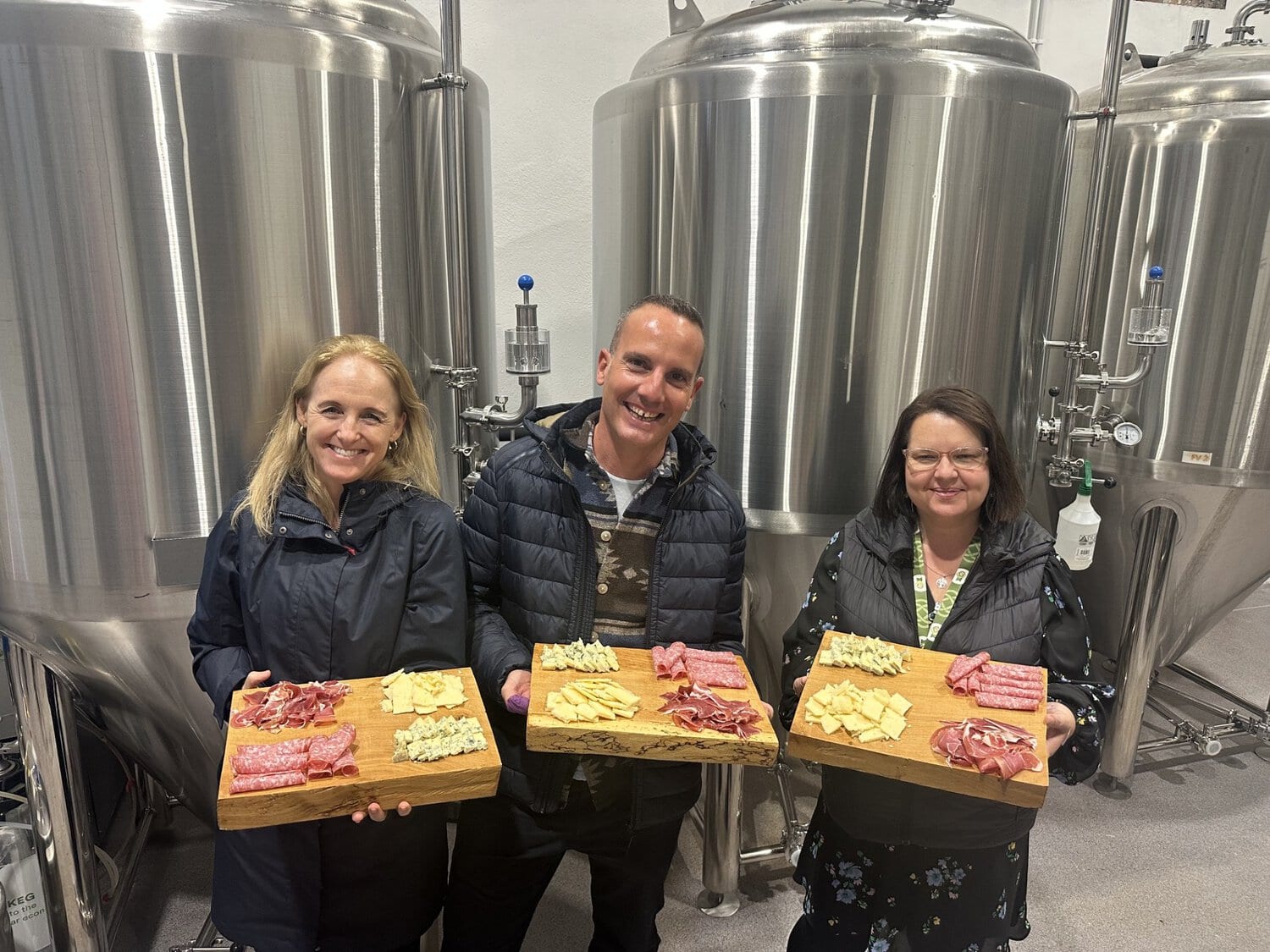 Drei Personen stehen vor großen Edelstahltanks in einer Brauerei und halten jeweils ein Holzbrett mit verschiedenen Käse- und Wurstspezialitäten in den Händen.