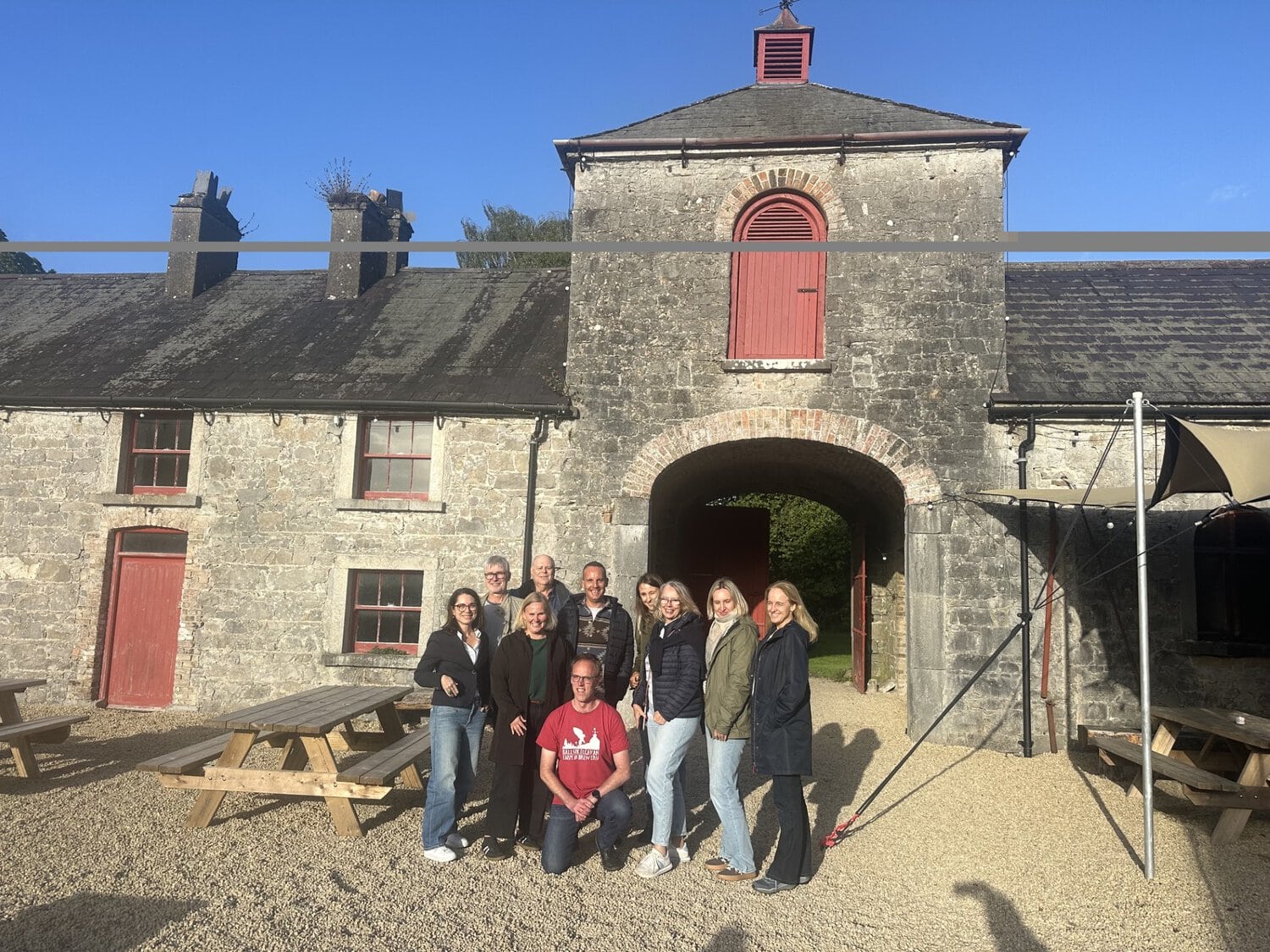 Eine Gruppe von neun Personen posiert vor einem historischen Steingebäude mit einem Torbogen und einem Turmaufbau auf einem geschotterten Innenhof.