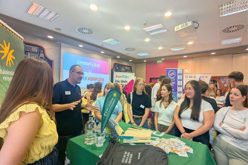 Ein Mann im schwarzen Poloshirt spricht vor einer Gruppe junger Erwachsener an einem Messestand mit grüner Tischdecke und verschiedenen Werbeartikeln.