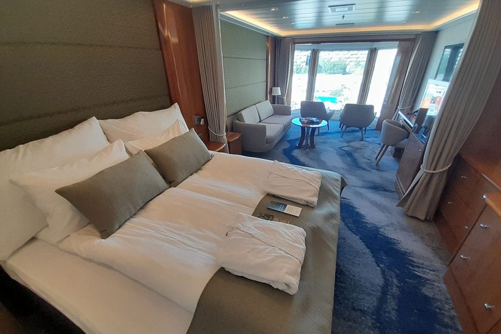 Blick in eine luxuriöse Schiffskabine mit großem Doppelbett, zwei Bademänteln, einer Sitzgruppe mit Sofa und Sesseln sowie einem Fenster mit Außenblick.