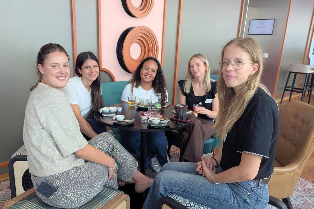 Fünf junge Frauen sitzen gemeinsam an einem runden schwarzen Tisch in einem modern eingerichteten Raum und blicken freundlich in die Kamera des Fotografen.