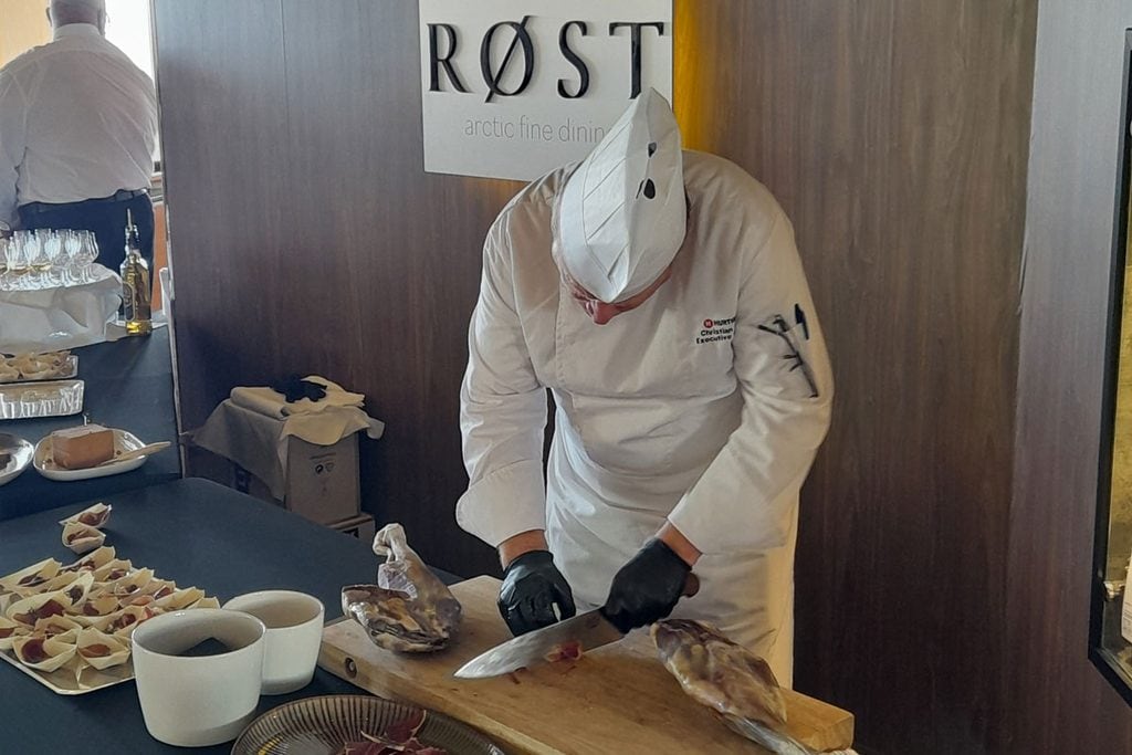 Ein Koch in weißer Uniform und Kochmütze schneidet mit einem großen Messer Schinken von einem Stück auf einem Holzbrett vor einem RØST-Wandschild.