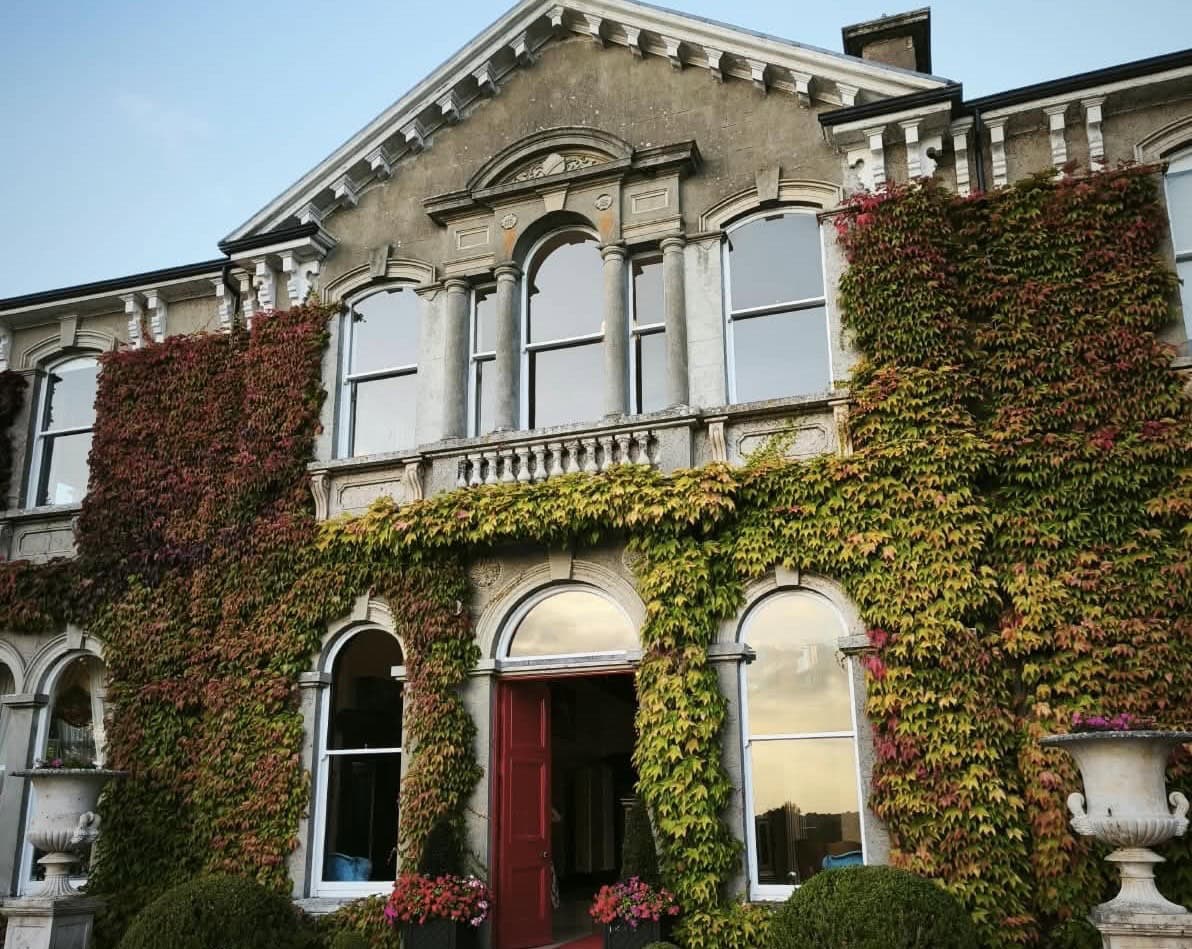 Die Fassade eines historischen Steingebäudes mit einer roten Eingangstür, das von dichtem, herbstlich verfärbtem Efeu und zwei Steinvasen umgeben ist.