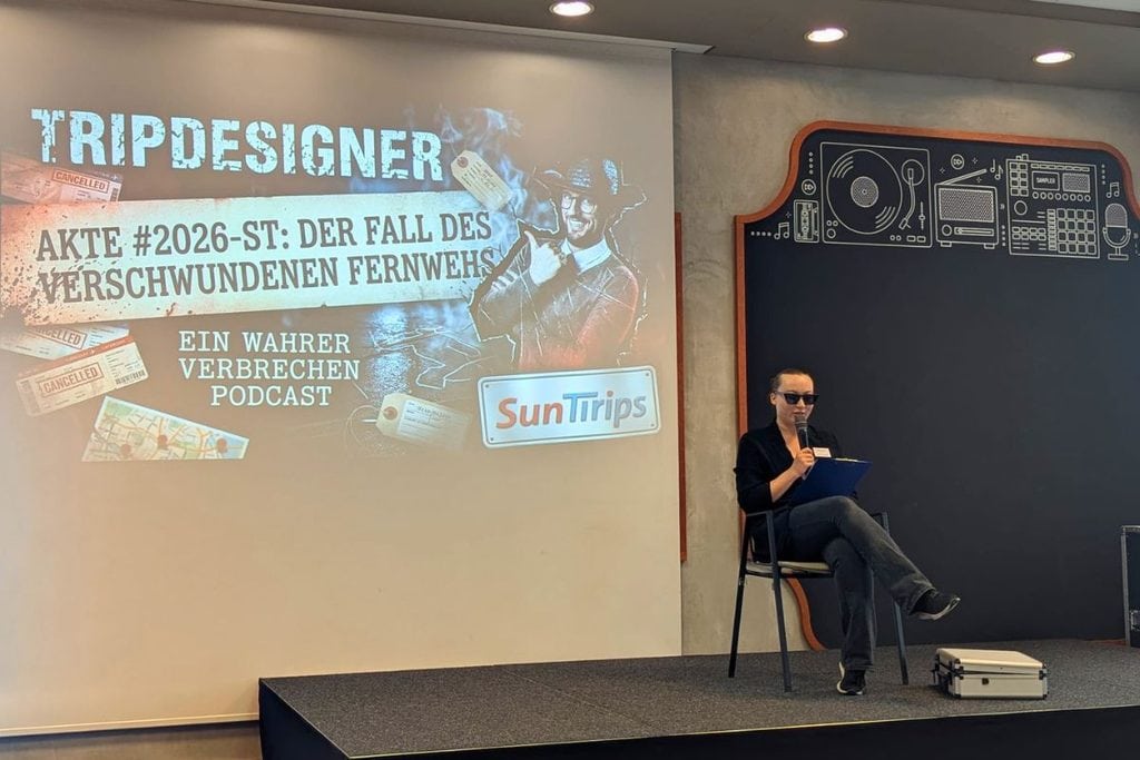 Eine Person mit Sonnenbrille sitzt auf einer Bühne vor einer großen Projektionsfläche mit dem Titel TRIPDESIGNER und hält ein Mikrofon in der Hand.