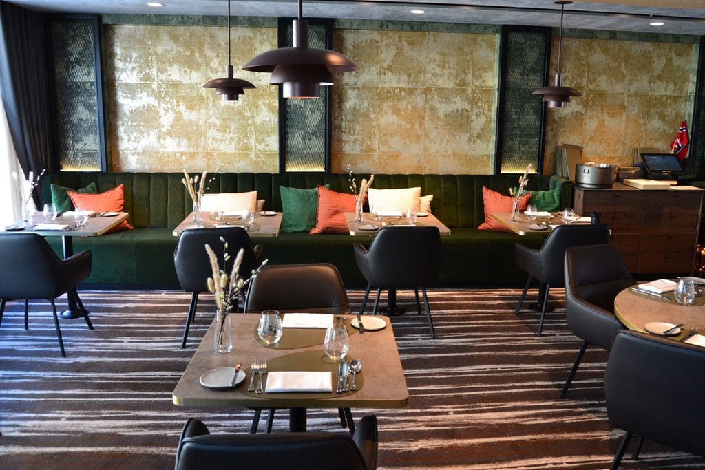 Ein Restaurantbereich mit einer langen grünen Sitzbank, gedeckten Tischen, dunklen Stühlen und einer strukturierten Wandgestaltung in Gold- und Brauntönen.