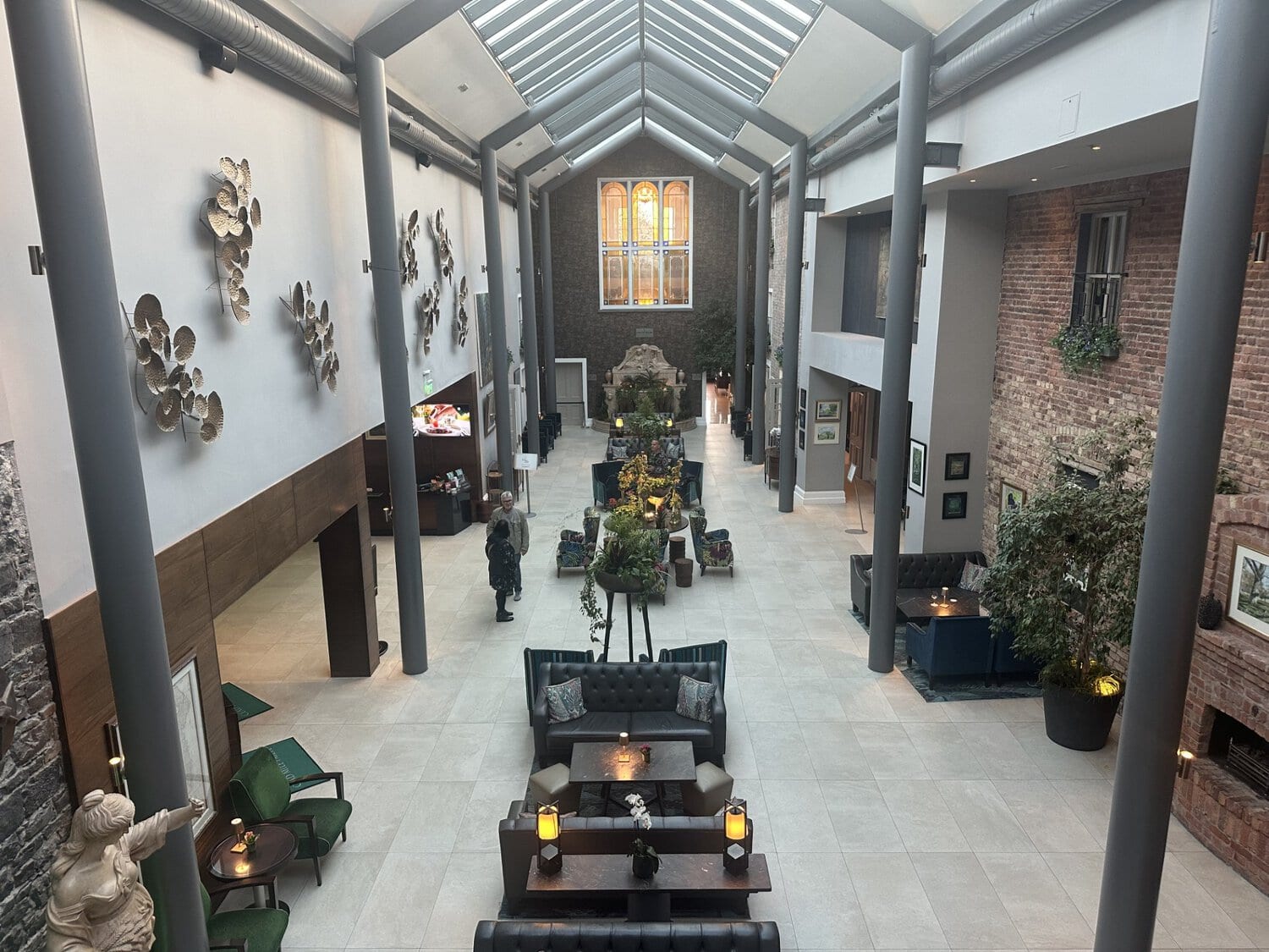 Blick von oben in eine hohe Hotellobby mit Glasdach, Säulen, Sitzgruppen aus Ledersofas und Sesseln sowie einer Wanddekoration aus metallischen Elementen.