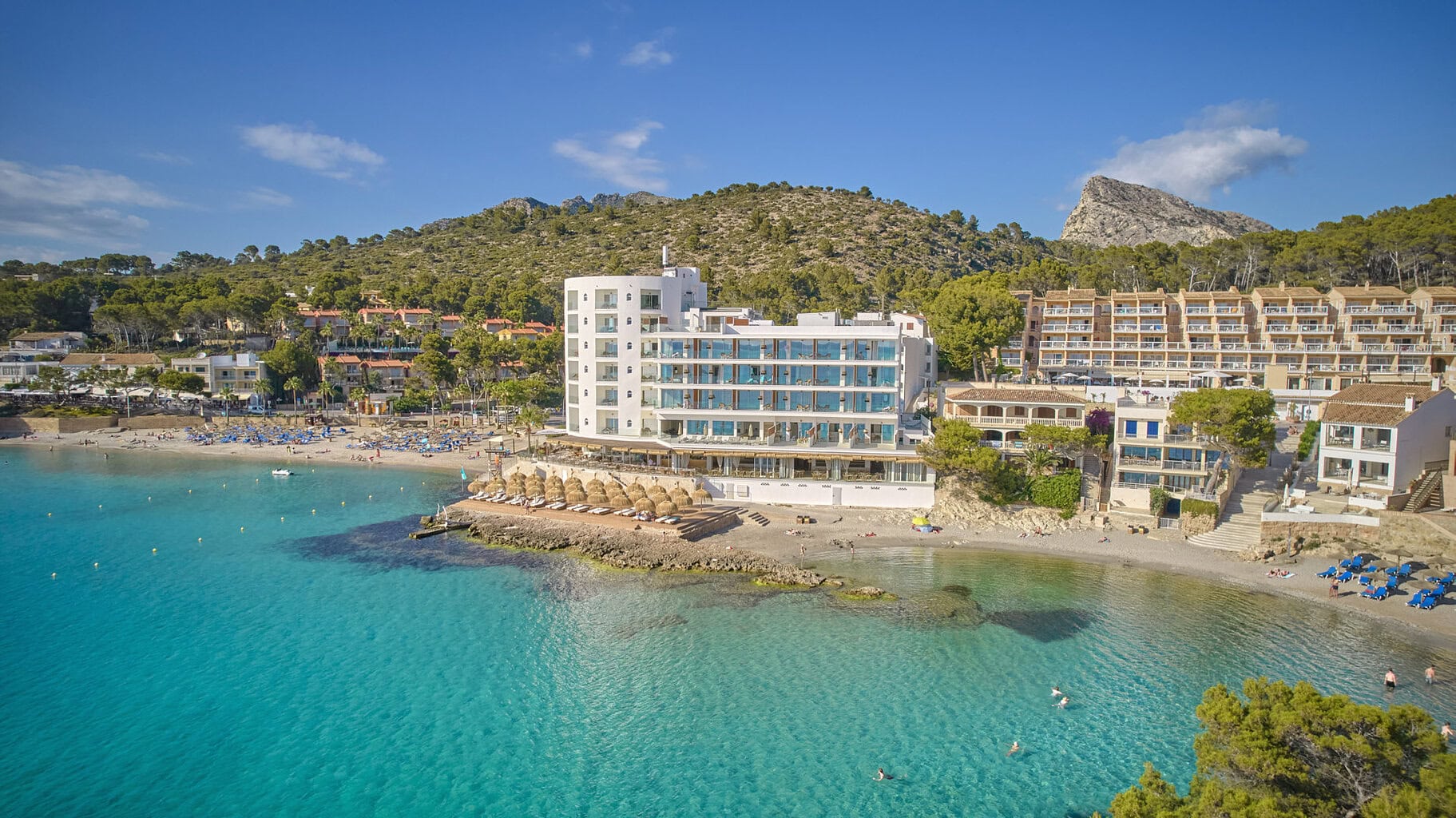 Luftaufnahme eines Hotels an einer Bucht mit türkisfarbenem Wasser, Sandstrand, Sonnenschirmen und einer hügeligen Landschaft im Hintergrund bei blauem Himmel.