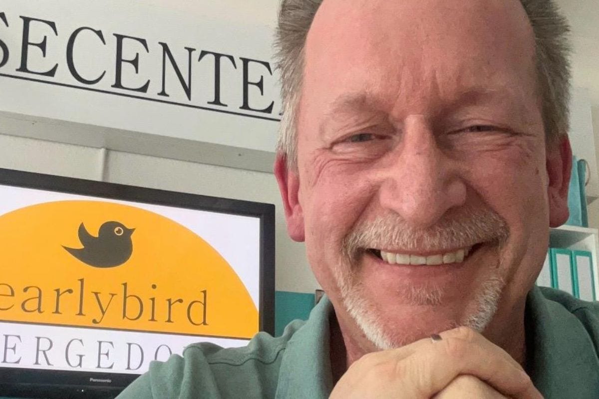 Ein lächelnder Mann mit Bart sitzt vor einem Bildschirm mit dem Logo von Earlybird. Im Hintergrund sind Aktenordner sichtbar.