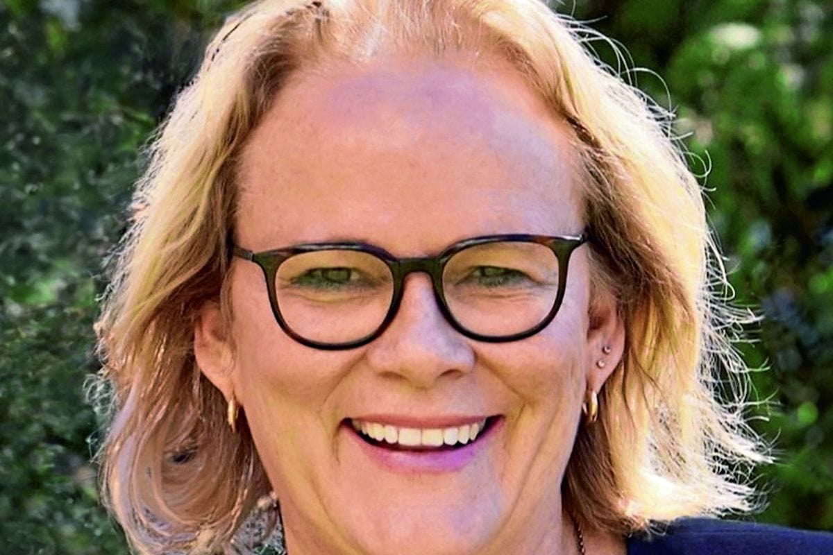 Nahaufnahme einer lächelnden Frau mit blondem, schulterlangem Haar, die eine dunkle Brille trägt und vor einem unscharfen, grünen Hintergrund posiert.